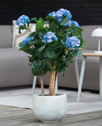 Een Kunstplant Hortensia UV 85 cm blauw met blauwe bloemen en groene bladeren staat op een houten vloer voor een grijze bank en een klein rond tafeltje met een witte lamp.