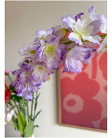 Close-up van de Kunstbloem Delphinium 105 cm paars, geschikt in een vaas, met op de achtergrond een roze en rood bloemenkunstwerk aan de muur.