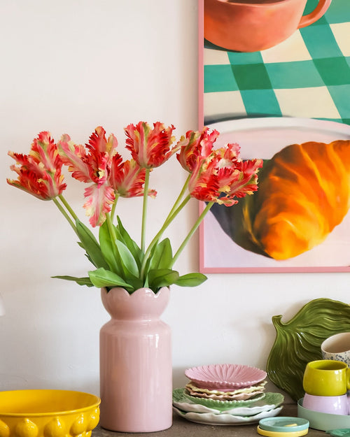 Su un tavolo con stoviglie colorate si trova un vaso rosa contenente il fiore artificiale Tulipano Parrocchetto Francese 76 cm rosso/giallo, mentre sullo sfondo si vede in parte un dipinto incorniciato di un croissant e una tazza.