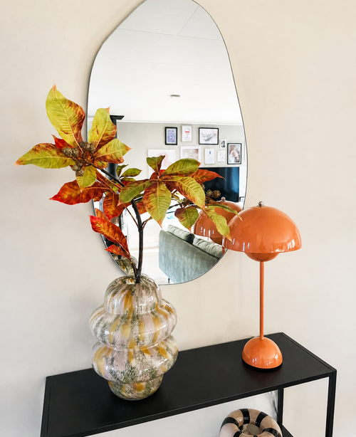 In uno specchio ondulato sopra una consolle nera si trovano un vaso di vetro con Kunsttak Chestnut 91 cm rosso/verde e una lampada a forma di fungo arancione. Lo specchio riflette un soggiorno con quadri murali incorniciati.