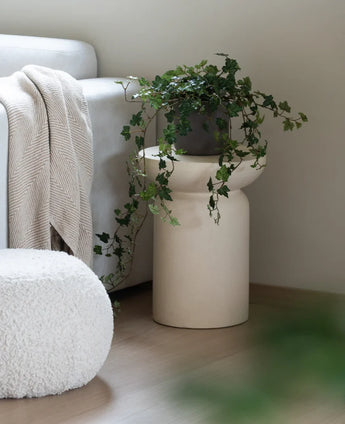 Een Kunst Klimop Deluxe Groen 90 cm staat in een beige vaas op een houten vloer naast een lichtgrijze bank waarop een crèmekleurige plaid ligt en een witte poef met structuur.