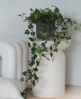Een Kunst Klimop Deluxe Groen, een hangende klimop van 90 cm in een grijze pot, staat op een modern, wit, sculpturaal voetstuk naast een opgevouwen witte deken en een wit kussen met structuur.