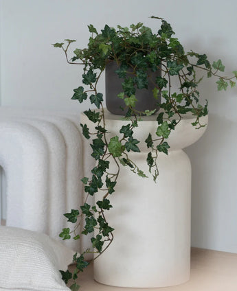 Een Kunst Klimop Deluxe Groen, een hangende klimop van 90 cm in een grijze pot, staat op een modern, wit, sculpturaal voetstuk naast een opgevouwen witte deken en een wit kussen met structuur.