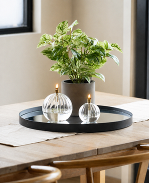 Una vite dorata artificiale verde-bianca in vaso UV 35 cm sta insieme a due lampade a olio in vetro su una piattaforma a specchio rotonda su un tavolo di legno, illuminato dalla luce naturale che filtra attraverso una finestra vicina in questo ambiente moderno e minimalista.
