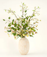 Een hoge vaas van beige keramiek bevat het Zijden kunstbloemen boeket Wild Apple Blossom XL, met groene bladertakken, kleine gele bloemen en roze bessen tegen een lichte achtergrond.