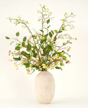 Un vaso alto in ceramica beige contiene il bouquet di fiori artificiali Silk Wild Apple Blossom XL, con rami frondosi verdi, piccoli fiori gialli e bacche rosa su uno sfondo chiaro.
