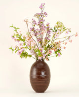 Een bruine vaas van keramiek toont het Zijden kunstbloemen boeket Lilac, een hoog en elegant zijden boeket met roze, paarse en lila bloemen met groene bladeren en dunne takken tegen een effen lichte achtergrond.