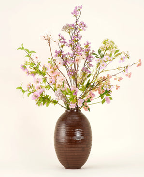 Un vaso in ceramica marrone mostra il bouquet di fiori artificiali Silk Lilac, un bouquet di seta alto ed elegante con fiori rosa, viola e lilla con foglie verdi e rami sottili su uno sfondo chiaro.