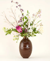 Een Zijden kunstbloemen boeket Serene Bloomfield, met een grote roze protea, witte rozen, paarse en witte bloesems en groen gebladerte, staat in een bruine geribbelde vaas tegen een effen lichte achtergrond.