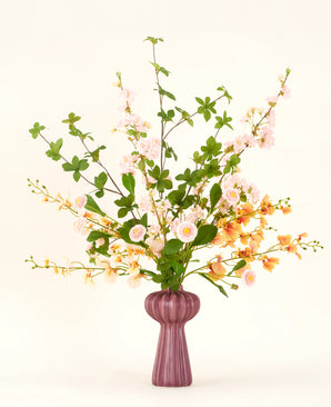 Un bouquet di fiori artificiali in seta Blossom Harmony è disposto in un vaso color malva con fiori rosa e pesca e foglie verdi, su un semplice sfondo chiaro per un'esposizione elegante.