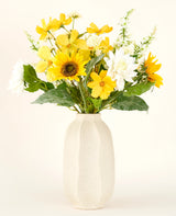 Het Zijden kunstbloemen boeket Sunny Whisper, met zonnebloemen, witte dahlia's en groene bladeren in een beige keramieken vaas, komt prachtig tot zijn recht tegen een lichte achtergrond.