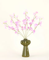 Een groene glazen vaas toont het Zijden kunstbloemen boeket Erwt roze met zachtgroen gebladerte, met verschillende lange stelen en tegen een effen gebroken witte achtergrond.
