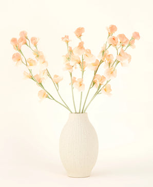 Il vaso in ceramica color crema contiene il bouquet di fiori artificiali Silk Pea Peach, con delicati fiori color pesca e rosa chiaro con lunghi steli verdi su uno sfondo chiaro e neutro.