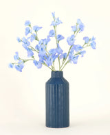 Een blauw geribbelde vaas van keramiek toont het Zijden kunstbloemen boeket Erwt blauw, met zijn delicate lichtblauwe kunstbloemen tegen een effen, lichte achtergrond.