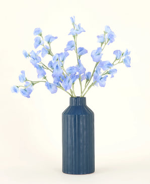 Un vaso in ceramica a coste blu mostra il bouquet di fiori artificiali in seta Pea Blue, con i suoi delicati fiori artificiali azzurri su uno sfondo semplice e chiaro.
