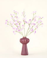 Het Zijden kunstbloemen boeket Erwt lila heeft lange stelen van delicate, lichtpaarse bloemen en groene bladeren in een vaas van mauve keramiek met verticale richels, tegen een effen, gebroken witte achtergrond.