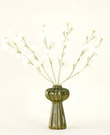 Een groene geribbelde glazen vaas bevat het Zijden kunstbloemen boeket Erwt crème met delicate witte zijden bloemen en slanke stelen, tegen een effen gebroken witte achtergrond.