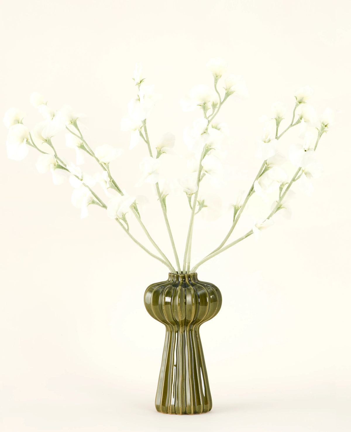 Un vaso di vetro verde a coste contiene il bouquet di fiori artificiali in seta Crema di piselli con delicati fiori di seta bianca e steli sottili, su un semplice sfondo bianco sporco.