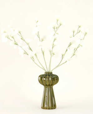 Un vaso di vetro verde a coste contiene il bouquet di fiori artificiali in seta Crema di piselli con delicati fiori di seta bianca e steli sottili, su un semplice sfondo bianco sporco.