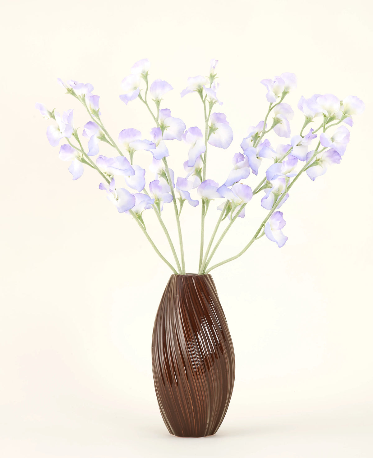 Un vaso marrone a coste contiene il bouquet di fiori di seta artificiale lavanda pisello, con petali delicati e steli verdi su un semplice sfondo bianco sporco per una sottile atmosfera da cottage.
