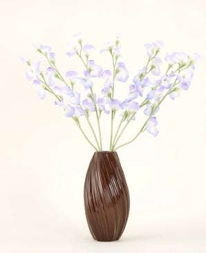 Un vaso marrone a coste contiene il bouquet di fiori di seta artificiale lavanda pisello, con petali delicati e steli verdi su un semplice sfondo bianco sporco per una sottile atmosfera da cottage.
