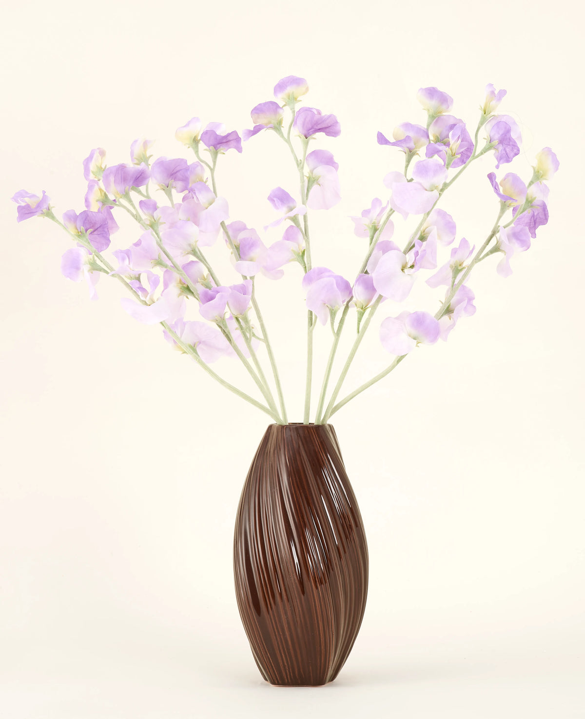 Un vaso marrone strutturato contiene il bouquet di fiori di seta artificiale Fiori di seta viola-viola chiaro di pisello con steli verdastri su uno sfondo semplice e chiaro.