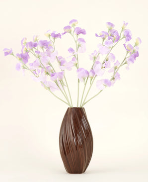 Un vaso marrone strutturato contiene il bouquet di fiori di seta artificiale Fiori di seta viola-viola chiaro di pisello con steli verdastri su uno sfondo semplice e chiaro.