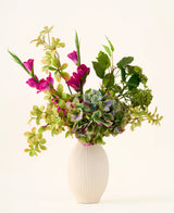 In een witte vaas met structuur staat een boeket van groene hortensia’s, bladrijke takken en felmagenta zijden bloemen tegen een effen lichte achtergrond, waardoor een opvallend ‘Cottage Garden’-boeket van zijden kunstbloemen ontstaat dat elke ruimte een blijvende charme geeft.