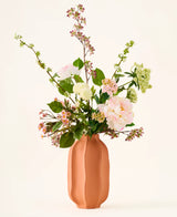 In un vaso di ceramica color pesca c'è un bouquet di fiori artificiali di seta rosa e bianca, il bouquet "Dreamy Meadow", con foglie e steli verdi, su uno sfondo semplice e chiaro.