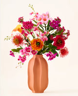 In un vaso color pesca si trova il bouquet "Tropical Sunset" di fiori artificiali di seta, composto da un vivace mix di fiori rosa, arancioni, gialli e rossi su uno sfondo semplice e chiaro.