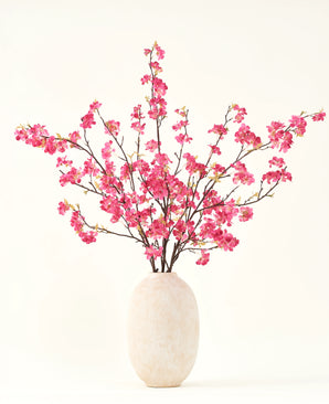 Il bouquet di fiori artificiali in seta Fiore di ciliegio fucsia 150 cm presenta vivaci steli di fiori di ciliegio fucsia con foglie verdi in un semplice vaso bianco, che completerà qualsiasi interno su uno sfondo chiaro.