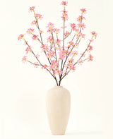 Een hoge vaas van beige keramiek bevat verschillende takken uit het Zijden kunstbloemen boeket Bloemen roze, met delicate roze bloesems en groene bladeren, tegen een effen witte achtergrond.