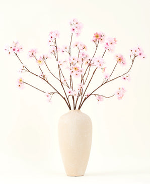 Il bouquet di fiori artificiali in seta Fiore di ciliegio rosa 114 cm ha rami lunghi e sottili con mazzi di fiori rosa chiaro in un vaso di ceramica beige, che formano un sorprendente contrasto con il semplice sfondo bianco.
