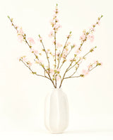Het Zijden kunstbloemen boeket Kersenbloesem lichtroze 94 cm heeft delicate roze kunstmatige kersenbloesems in een witte keramieken vaas, die elegantie toevoegt tegen een effen lichte achtergrond.