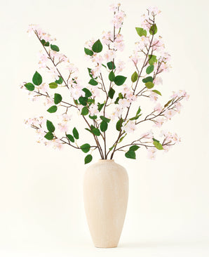 Un vaso in ceramica beige contiene diversi rami lunghi con il bouquet di fiori artificiali Silk di fiori di pesco rosa e foglie verdi, su uno sfondo bianco semplice, perfetto per un interno floreale artificiale.