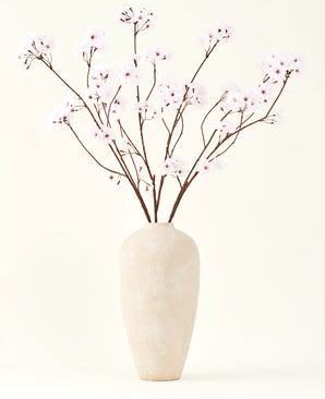 Il bouquet di fiori artificiali in seta Fiore di ciliegio rosa chiaro 114 cm si trova in un alto vaso di ceramica beige con rami lunghi e sottili e delicati fiori rosa chiaro, creando un sereno accento floreale artificiale su uno sfondo semplice.