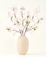 Het Zijden kunstbloemen boeket Kersenbloesem blush 119 cm toont delicate, lichtroze kersenbloesem met groene bladeren in een beige vaas, ideaal als kunstbloemen decoratie tegen een lichte achtergrond.