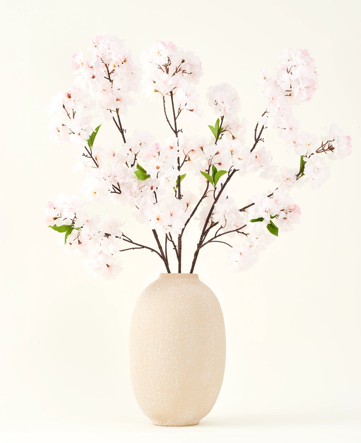 Il bouquet di fiori artificiali in seta Cherry Blossom Blush 119 cm mostra delicati fiori di ciliegio rosa chiaro con foglie verdi in un vaso beige, ideali come decorazione floreale artificiale su uno sfondo chiaro.
