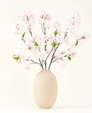 Il bouquet di fiori artificiali in seta Cherry Blossom Blush 119 cm mostra delicati fiori di ciliegio rosa chiaro con foglie verdi in un vaso beige, ideali come decorazione floreale artificiale su uno sfondo chiaro.