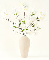 Het Zijden kunstbloemen boeket Kersenbloesem crème 119 cm heeft elegante bruine takken met zacht witte nepbloesem en groene bladeren, weergegeven in een eenvoudige beige vaas tegen een effen lichte achtergrond.
