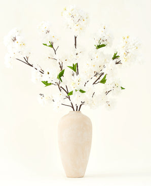 Il bouquet di fiori artificiali in seta Fiore di ciliegio crema 119 cm ha eleganti rami marroni con morbidi fiori finti bianchi e foglie verdi, esposti in un semplice vaso beige su uno sfondo chiaro.