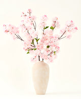 De Zijden kunstbloemen boeket Kersenbloesem roze 119 cm, gerangschikt in een beige keramische vaas met delicate groene bladeren, creëert een sereen interieuraccent tegen een eenvoudige lichte achtergrond.
