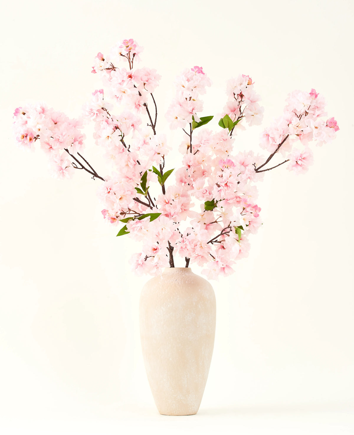 Il bouquet di fiori artificiali Silk Fiore di ciliegio rosa 119 cm, disposto in un vaso di ceramica beige con delicate foglie verdi, crea un sereno accento interno su un semplice sfondo chiaro.