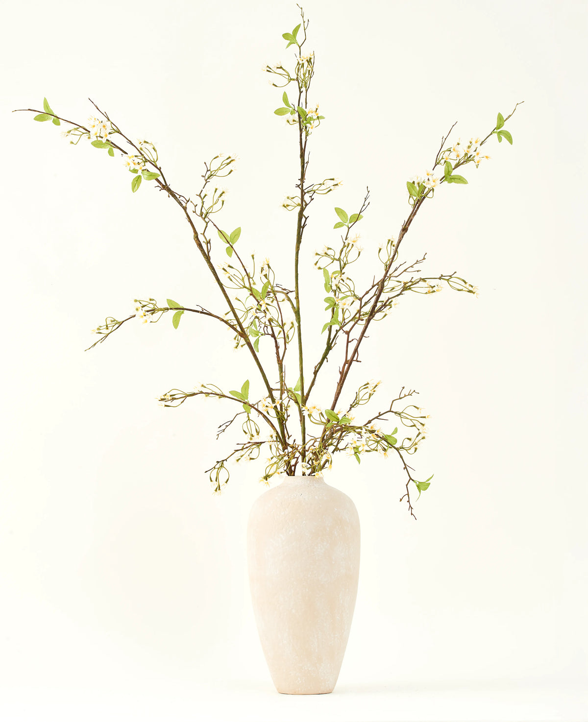 Un vaso alto in ceramica color crema contiene il bouquet di fiori artificiali in seta Verbena crema con lunghi rami, piccole foglie verdi e delicati fiori bianchi, splendidamente disposti su uno sfondo chiaro e semplice.