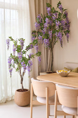 Twee Kunstplant Blauweregen van 150 cm, met hun overhangende paarse bloemen, staan naast een houten eettafel met lichte stoelen. Zacht natuurlijk licht valt door de doorschijnende gordijnen achter de planten naar binnen.