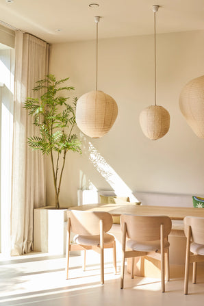 Una zona pranzo luminosa e minimalista è arredata con sedie in legno chiaro, un tavolo, tre lampade a sospensione in carta, la pianta di Mahonia artificiale di 210 cm in vaso e tende dal pavimento al soffitto vicino a una grande finestra che lascia entrare la luce del sole.