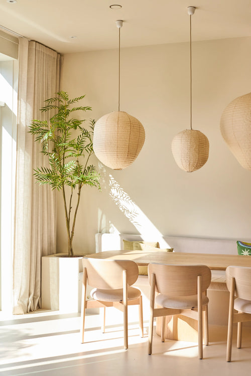 Una zona pranzo luminosa e minimalista è arredata con sedie in legno chiaro, un tavolo, tre lampade a sospensione in carta, la pianta di Mahonia artificiale di 210 cm in vaso e tende dal pavimento al soffitto vicino a una grande finestra che lascia entrare la luce del sole.