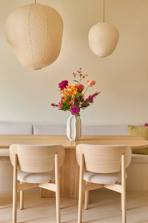Een minimalistische eethoek met een tafel en stoelen van licht hout, een beige bank, twee grote papieren lantaarns en een witte vaas met het boeket ‘Colourful Svenski’ van Zijden kunstbloemen als levendig middelpunt. De ruimte is ingericht in zachte, neutrale tinten, wat zorgt voor een gezellige en uitnodigende sfeer.