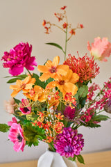 Een levendig boeket kunstbloemen van Zijden, ‘Magnus di Lambo’, bestaande uit een mix van oranje, roze en magenta kunstbloemen, is in een witte vaas geschikt tegen een neutrale achtergrond.