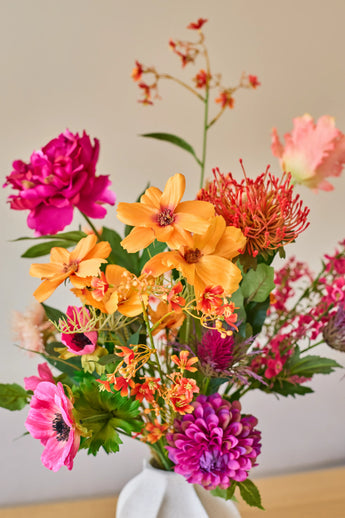 Een levendig boeket kunstbloemen van Zijden, ‘Magnus di Lambo’, bestaande uit een mix van oranje, roze en magenta kunstbloemen, is in een witte vaas geschikt tegen een neutrale achtergrond.